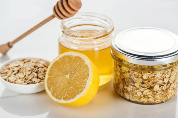 Enfoque selectivo de frascos de vidrio con miel y copos de avena, limón y palo de miel en la superficie blanca - foto de stock