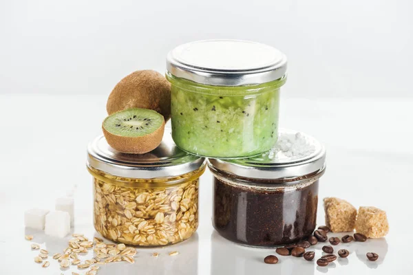 Glass jars with homemade cosmetics, kiwi and brown sugar cubes on white surface — Stock Photo