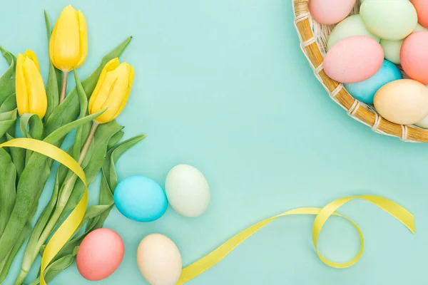 Top view of yellow tulips with ribbon and easter eggs in wicker plate isolated on blue — Stock Photo