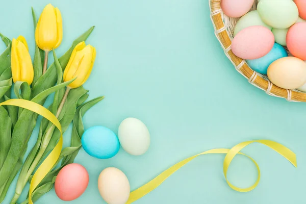 Vue de dessus des tulipes jaunes avec ruban et oeufs de Pâques dans une assiette en osier isolée sur bleu — Photo de stock