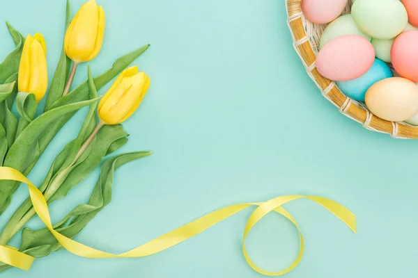 Vue de dessus des tulipes jaunes avec ruban et oeufs de Pâques dans une assiette en osier isolée sur bleu — Photo de stock