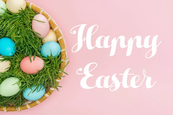 Top view of painted eggs in wicker plate with grass isolated on pink with Happy easter lettering — Stock Photo