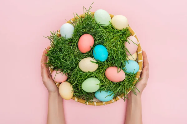 Vista recortada de la mujer con coloridos huevos de Pascua en plato de mimbre con hierba aislada en rosa - foto de stock