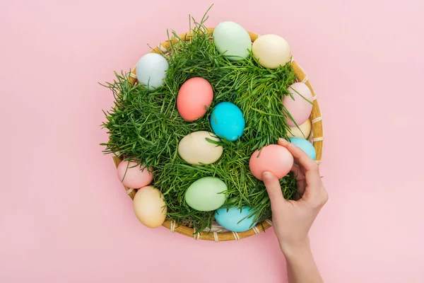 Vue recadrée de la main femelle avec des œufs de Pâques pastel dans une assiette en osier avec de l'herbe isolée sur rose — Photo de stock