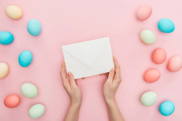 Vue partielle de la femme tenant enveloppe isolée sur rose avec des œufs de Pâques pastel — Photo de stock