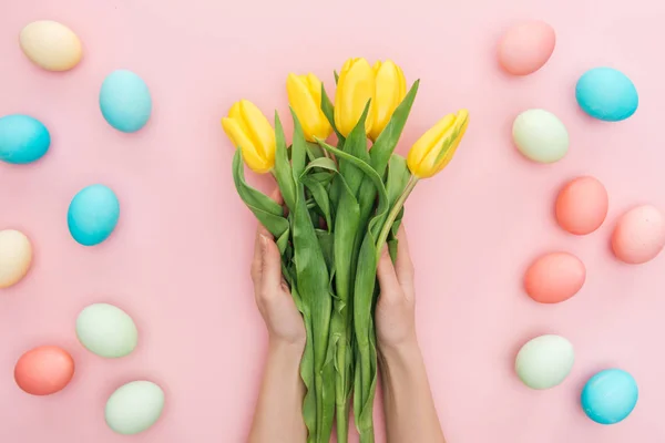 Vista cortada de mãos femininas com tulipas amarelas isoladas em rosa com ovos de páscoa — Fotografia de Stock