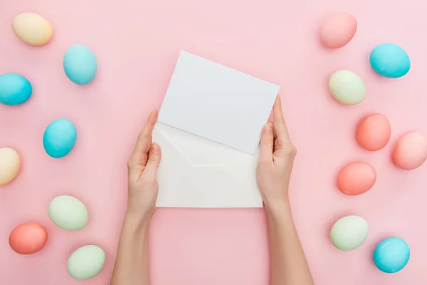 Abgeschnittene Ansicht weiblicher Hände mit Grußkarte und Umschlag isoliert auf rosa mit pastellfarbenen Ostereiern — Stockfoto