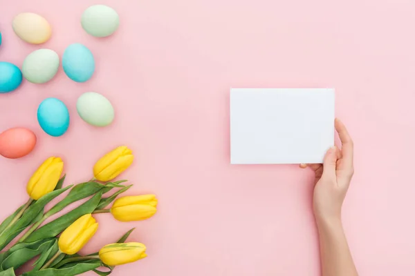 Vista recortada de la mano femenina con tarjeta vacía aislada en rosa con tulipanes amarillos y huevos de Pascua — Stock Photo