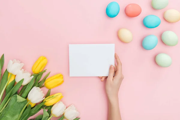 Vista recortada de las manos femeninas con tarjeta de felicitación aislada en rosa con tulipanes y huevos de Pascua - foto de stock