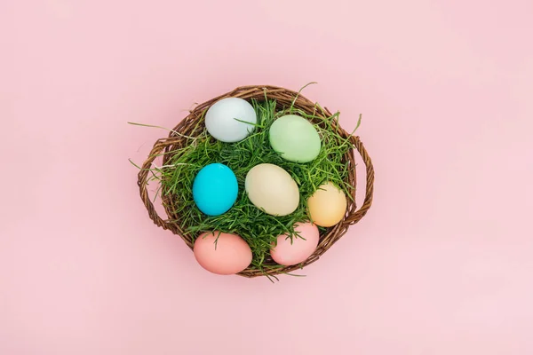 Vista superior de los huevos de Pascua en canasta de mimbre con hierba aislada en rosa - foto de stock