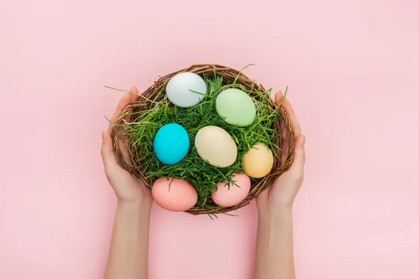 Vista recortada de manos femeninas sosteniendo plato de mimbre con hierba y huevos de Pascua aislados en rosa - foto de stock