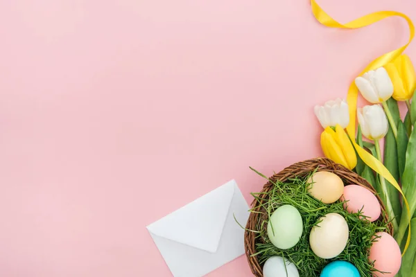 Vue du dessus de l'enveloppe, des fleurs de tulipes et des œufs de Pâques dans une assiette en osier isolée sur rose avec espace de copie — Photo de stock