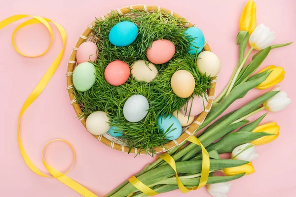 Vue du dessus des œufs de Pâques dans une assiette en osier avec herbe isolée sur rose avec des fleurs de tulipe — Photo de stock