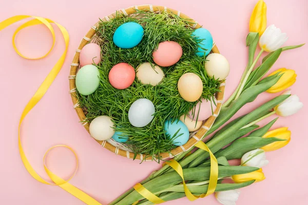 Vista superior de huevos de Pascua en plato de mimbre con hierba aislada en rosa con flores de tulipán - foto de stock