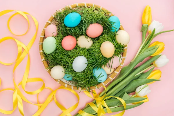 Vue du dessus des œufs de Pâques dans une assiette en osier avec herbe isolée sur rose avec des tulipes — Photo de stock