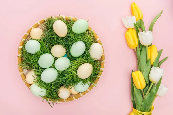 Vue du dessus des œufs de Pâques dans une assiette en osier avec herbe isolée sur rose avec des tulipes — Photo de stock