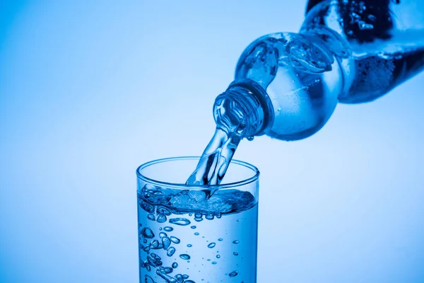 Agua que vierte de la botella de plástico en vidrio sobre fondo azul con espacio de copia - foto de stock