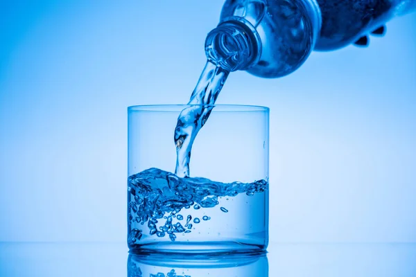 Vista recortada del hombre vertiendo agua de la botella de plástico en vidrio sobre fondo azul - foto de stock