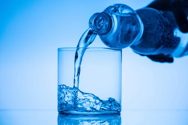 Обрезанный вид человека, наливающего воду из пластиковой бутылки в стекло на синем фоне — стоковое фото