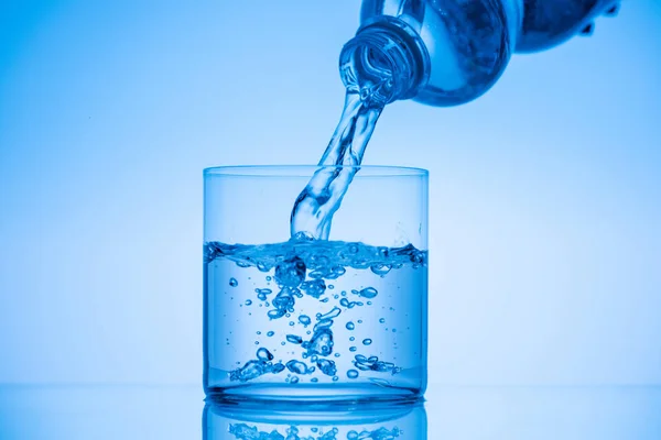 Water pouring from plastic bottle in drinking glass on blue background — Stock Photo