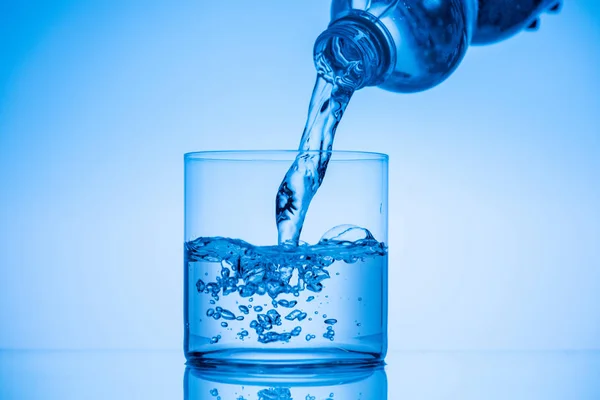 Acqua versando dalla bottiglia di plastica in vetro da bere su sfondo blu — Foto stock