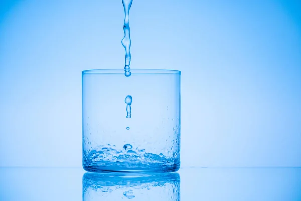 Imagem tonificada de água derramando em vidro vazio sobre fundo azul — Fotografia de Stock