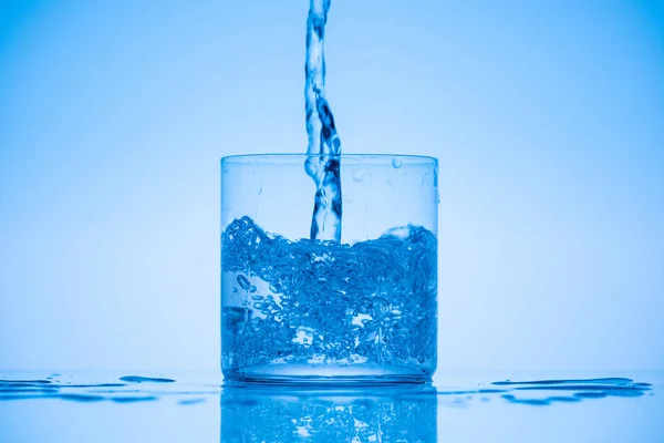 Тоноване зображення води, що ллється у питному склі на синьому фоні з бризками — Stock Photo