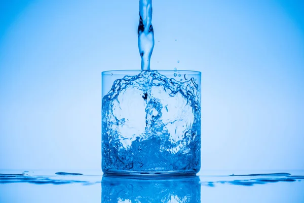 Toned image of water pouring in drinking glass on blue background with splashes — Stock Photo