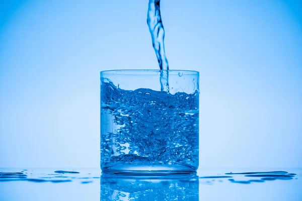 Toned image of water pouring in glass on blue background with splashes and copy space — Stock Photo