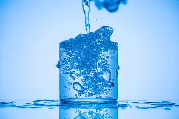 Immagine tonica di acqua versando in pieno vetro su sfondo blu con spruzzi — Foto stock