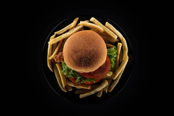 Vista superior de batatas fritas saborosas e hambúrguer de frango na placa isolada em preto — Fotografia de Stock