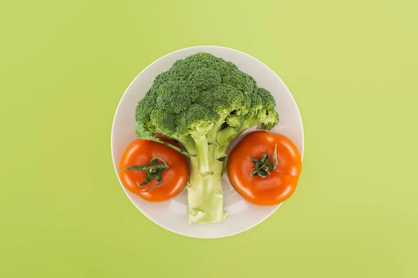 Top view of organic ripe broccoli near red tomatoes on white plate isolated on green — Stock Photo