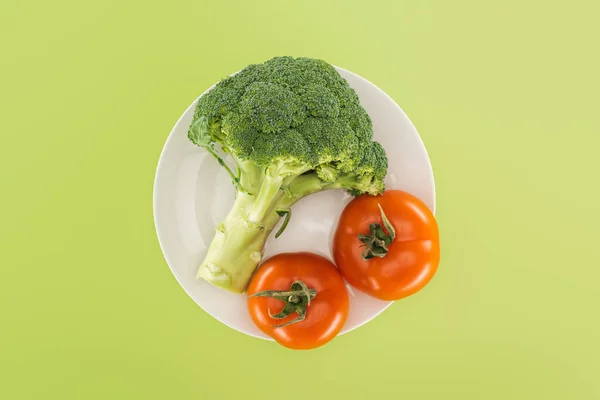 Top view of organic broccoli near red tomatoes on white plate isolated on green — Stock Photo