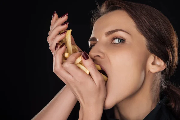 Mujer comiendo papas fritas mientras mira la cámara aislada en negro - foto de stock