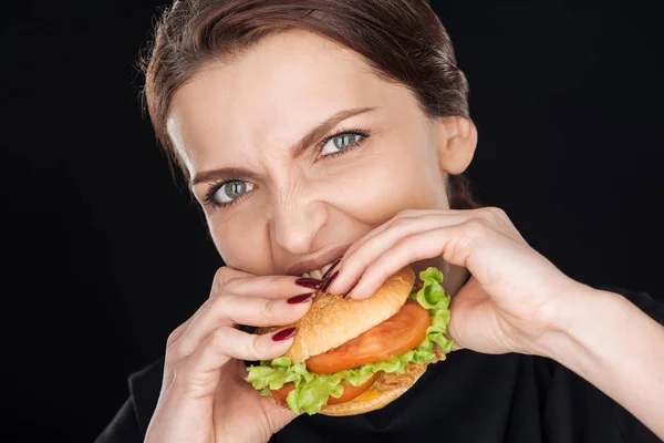 Раздраженная женщина ест вкусный куриный бургер, глядя на камеру, изолированную на черном — стоковое фото
