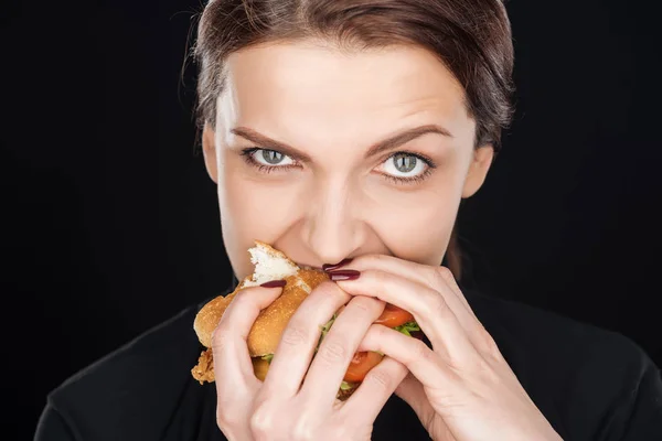 Hermosa mujer comiendo sabrosa hamburguesa de pollo mientras mira la cámara aislada en negro - foto de stock