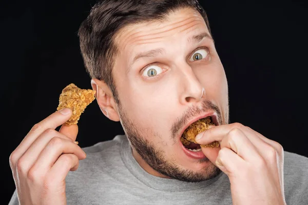 Uomo emotivo mangiare gustose pepite di pollo mentre guardando la fotocamera isolata su nero — Foto stock