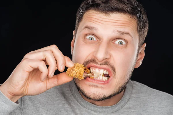 Emotional man eating crispy chicken nugget while looking at camera isolated on black — Stock Photo