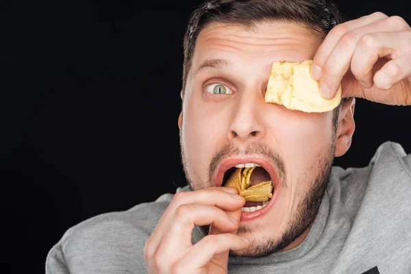 Homem cobrindo o olho com chip enquanto comia isolado em preto — Fotografia de Stock