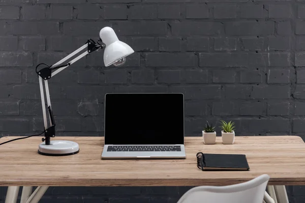 Modern workplace with laptop with blank screen, plants, lamp and notebook on wooden table near black brick wall — Stock Photo