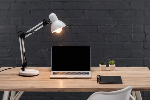 Local de trabalho moderno com laptop com tela em branco, plantas, lâmpada brilhante e notebook na mesa de madeira perto da parede de tijolo preto — Fotografia de Stock