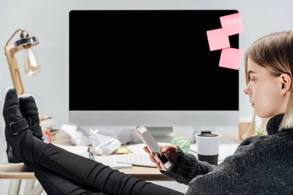 Selektiver Fokus des Mädchens, das am chaotischen Arbeitsplatz sitzt, die Beine auf dem Schreibtisch hat und das Smartphone isoliert auf grau setzt — Stockfoto