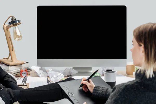 Girl sitting at messy workplace with legs on desk and using graphic tablet isolated on grey — Stock Photo