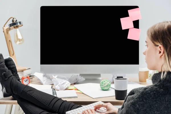 Girl sitting at messy workplace with legs on desk and typing on computer keyboard isolated on grey — Stock Photo