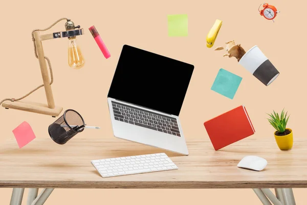 Laptop with blank screen, thermomug with coffee splash and stationery levitating in air above wooden desk isolated on beige — Stock Photo