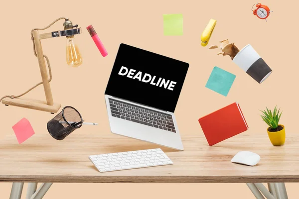 Laptop with deadline lettering on screen, thermomug with coffee splash and stationery levitating in air above wooden desk isolated on beige — Stock Photo