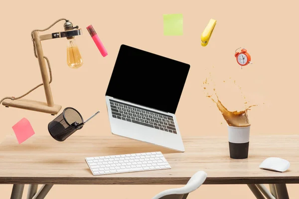 Laptop with blank screen, lamp, empty sticky notes and stationery levitating in air above workplace with thermomug with coffee splash  on table isolated on beige — Stock Photo