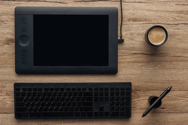 Vista superior de la tableta gráfica negra, pluma gráfica, teclado inalámbrico de la computadora y taza con café en la mesa de madera - foto de stock