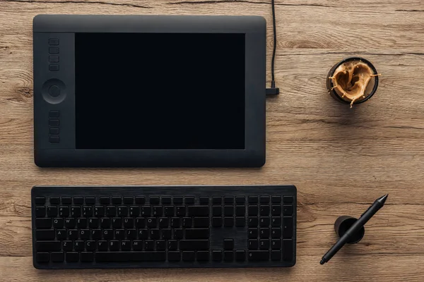 Vista superior de la tableta gráfica negro, pluma gráfica, teclado inalámbrico de la computadora y la taza con el café salpicadura en la mesa de madera - foto de stock