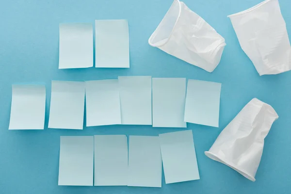 Top view of white plastic and crumpled cups and empty sticky notes on blue background — Stock Photo